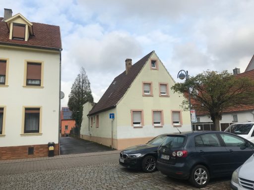 Zwei Häuser auf einer Straße, eines hellgrün, das andere beige, mit parkenden Autos.