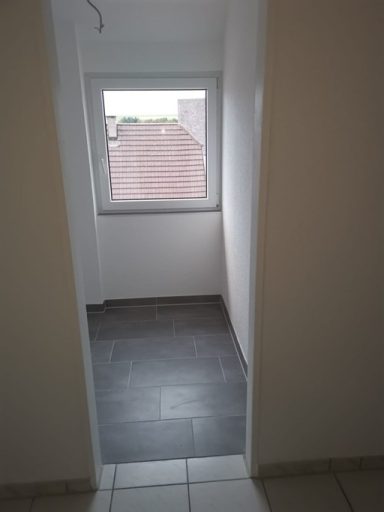 Heller Raum mit Fenster und grauem Fliesenboden, Blick auf ein Dach.