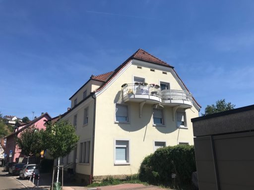 Helles, dreigeschossiges Gebäude mit Balkon, umgeben von Bäumen und Nachbarhäusern.