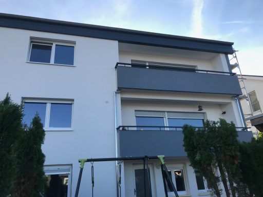 Modernes zweigeschossiges Gebäude mit zwei Balkonen und grünen Sträuchern im Vordergrund.