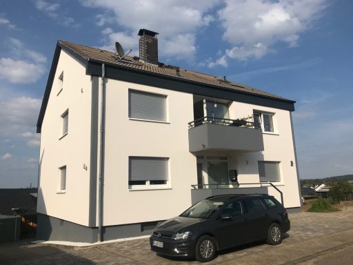 Helles zweigeschossiges Wohnhaus mit Balkon und schwarzem Auto davor.