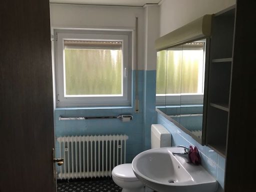 Kleines Badezimmer mit Waschbecken, Toilette und Fenster, blaue Wände und Heizkörper.