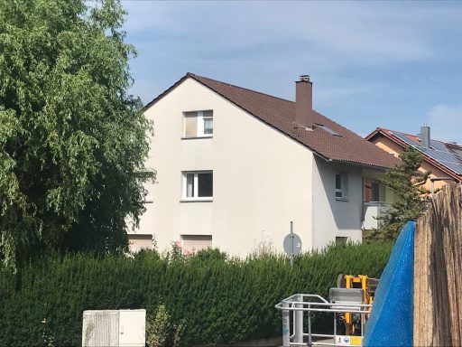 Helle Wohnhausfassade mit einem grauen Dach und angrenzender grüner Hecke.